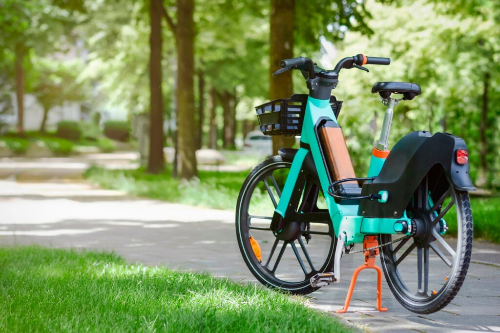 Turquoise e-bicycle with basket parked on a paved path in a leafy park.