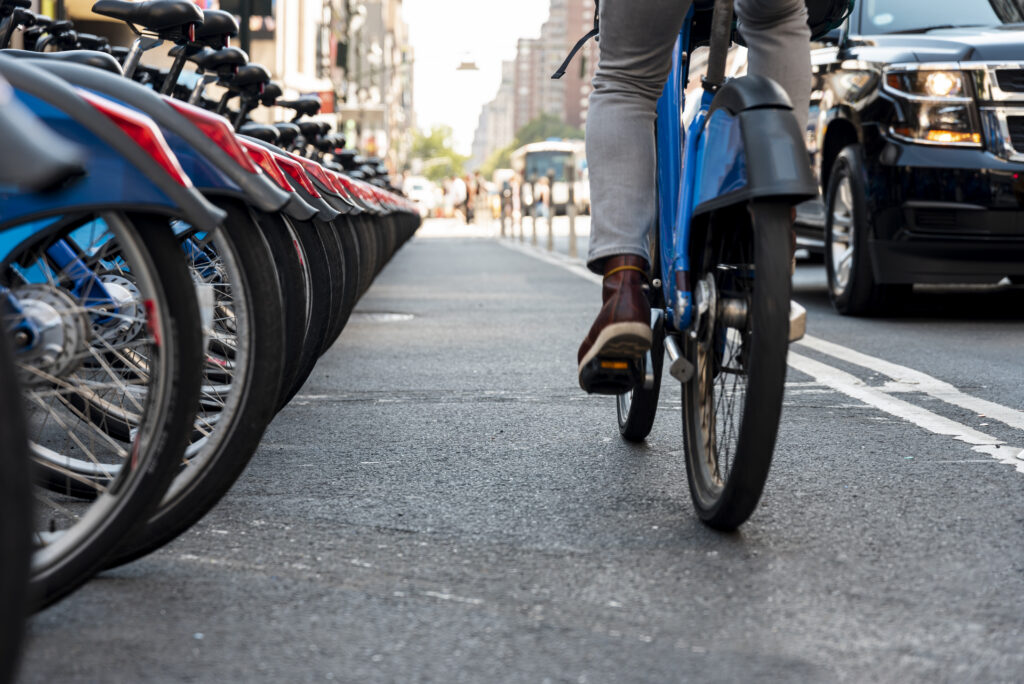 Close-up view of a person riding a blue e-bike on a city street next to a row of parked bicycles, with cars and pedestrians visible in the background.